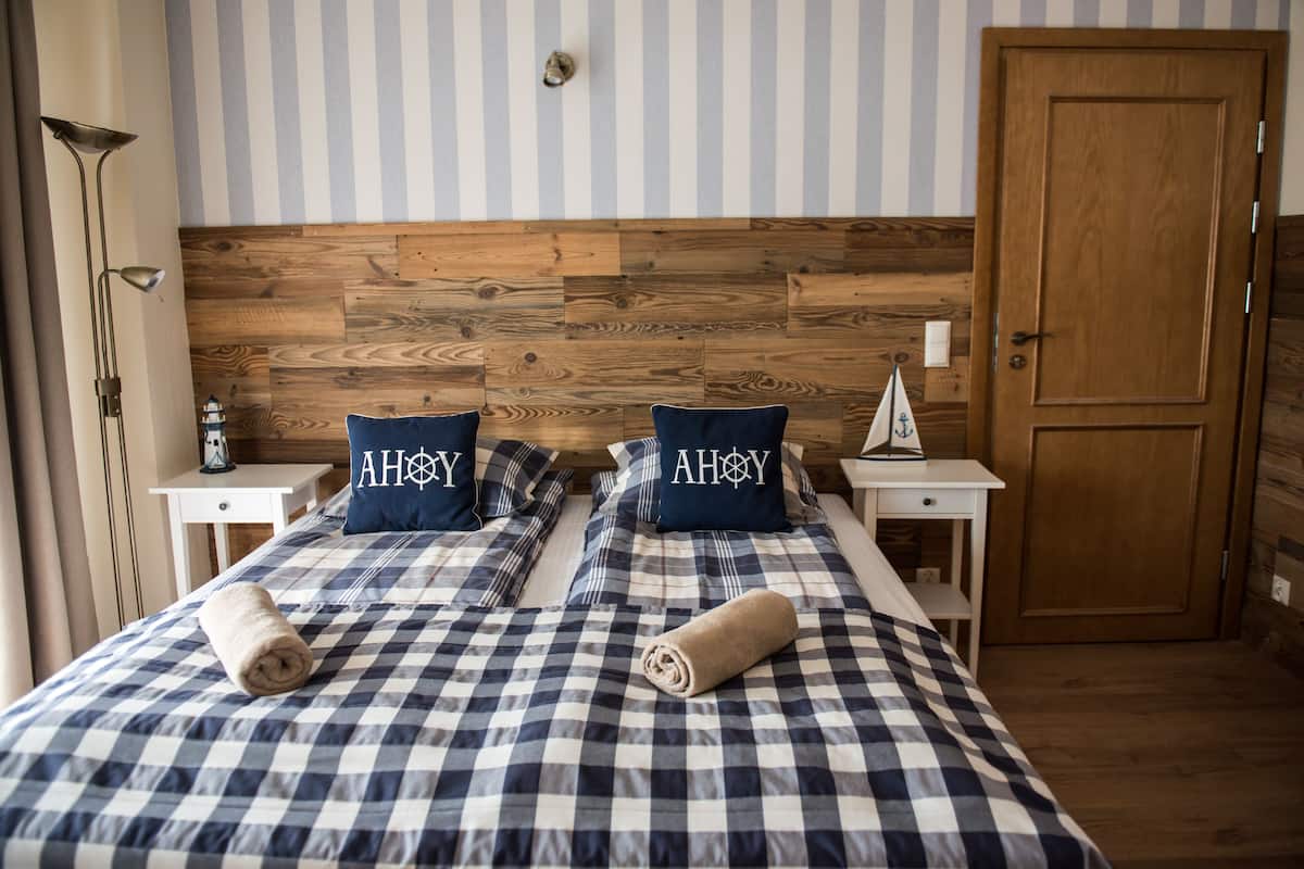 A hotel room with a checkered bedspread, two pillows with the text "AHOY," a wooden headboard, a nightstand with a lamp, and a wooden door.