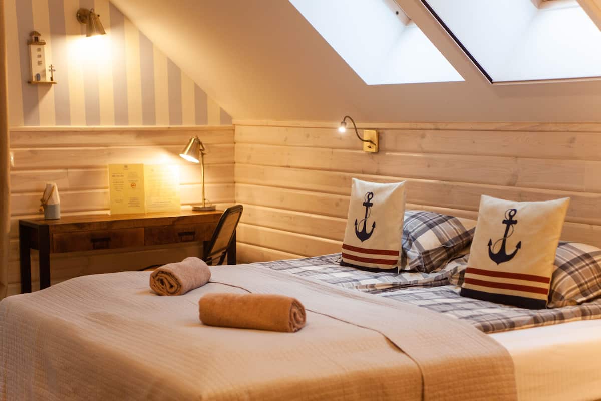 A cozy bedroom with a sloped ceiling, wooden walls, a desk with a lamp, and a bed with nautical-themed pillows.