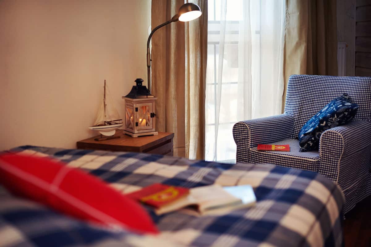A bedroom with a checkered bedspread, a red pillow, a small table with a lantern, and a checkered armchair with a blue pillow.