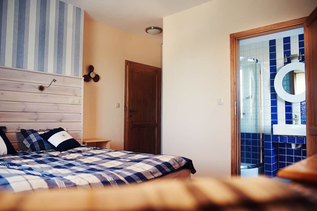 A bedroom with a wooden headboard, a checkered bedspread, a wooden door, and a bathroom with blue tiles and a round mirror.