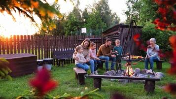 A group of people gathered around a campfire in an outdoor setting with a wooden fence and trees in the background.