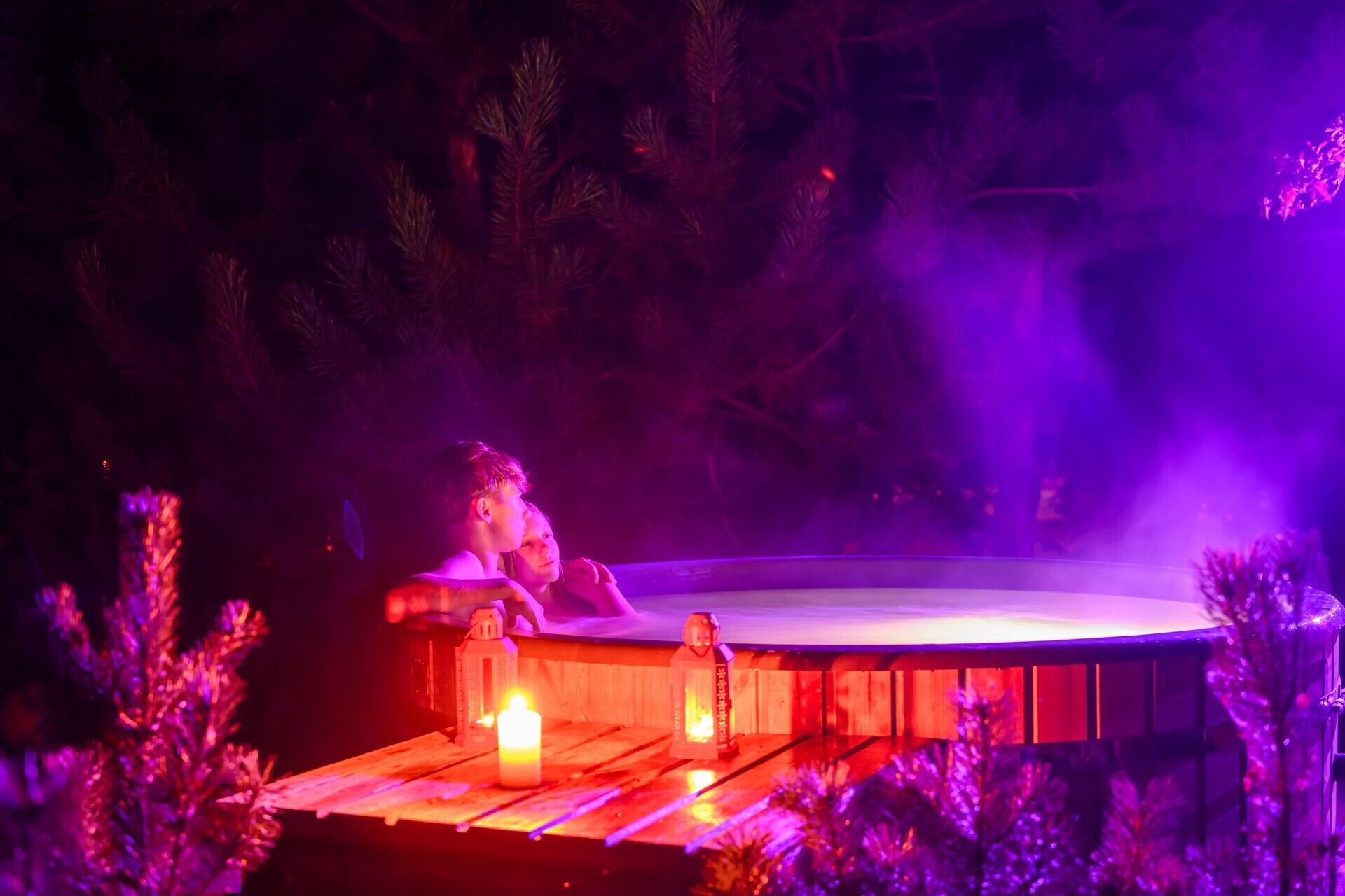 A person relaxing in a hot tub at night with purple lighting and candles.