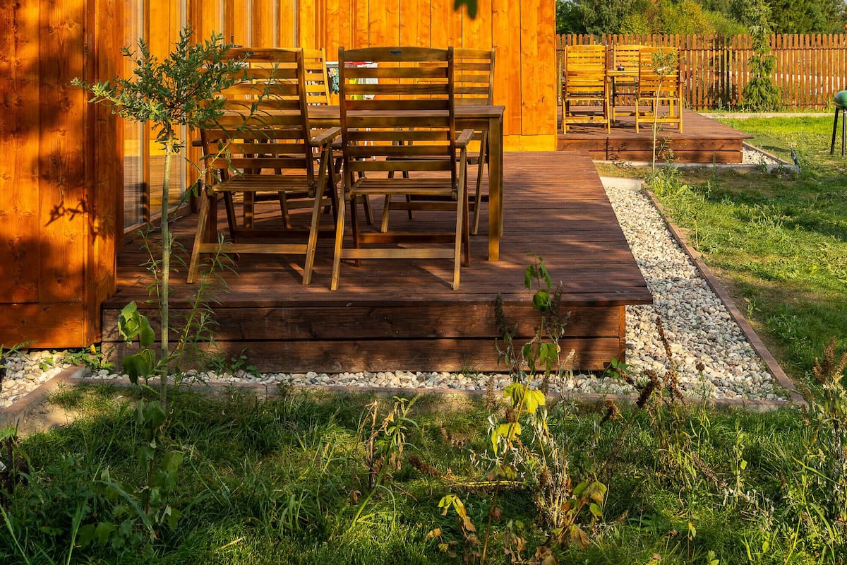 A wooden cabin with a red roof, a porch with wooden benches, and a gravel pathway.