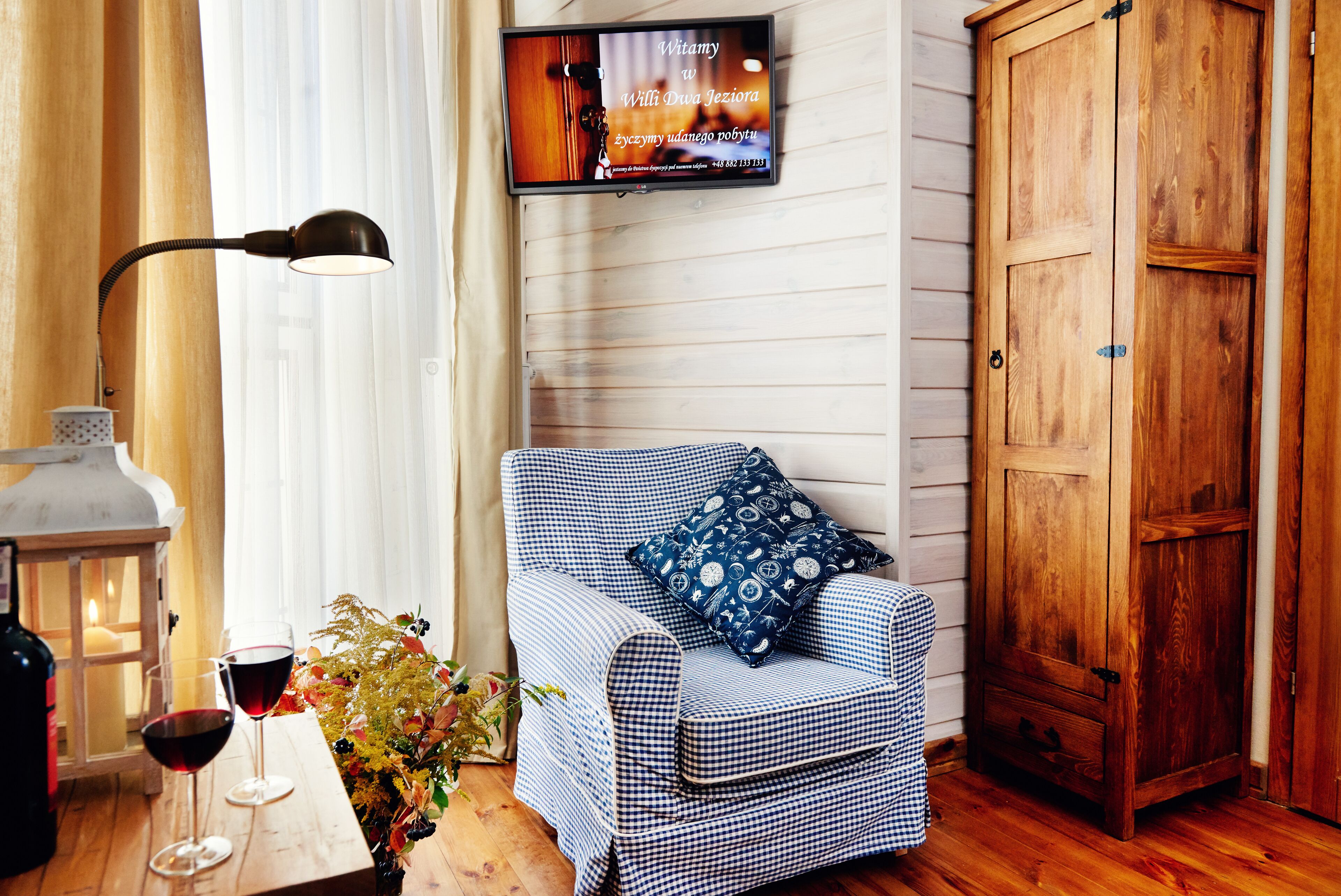 Una habitación acogedora con un sofá estampado de cuadros, un armario de madera, un televisor fijado a la pared y una mesa con una botella y vasos.
