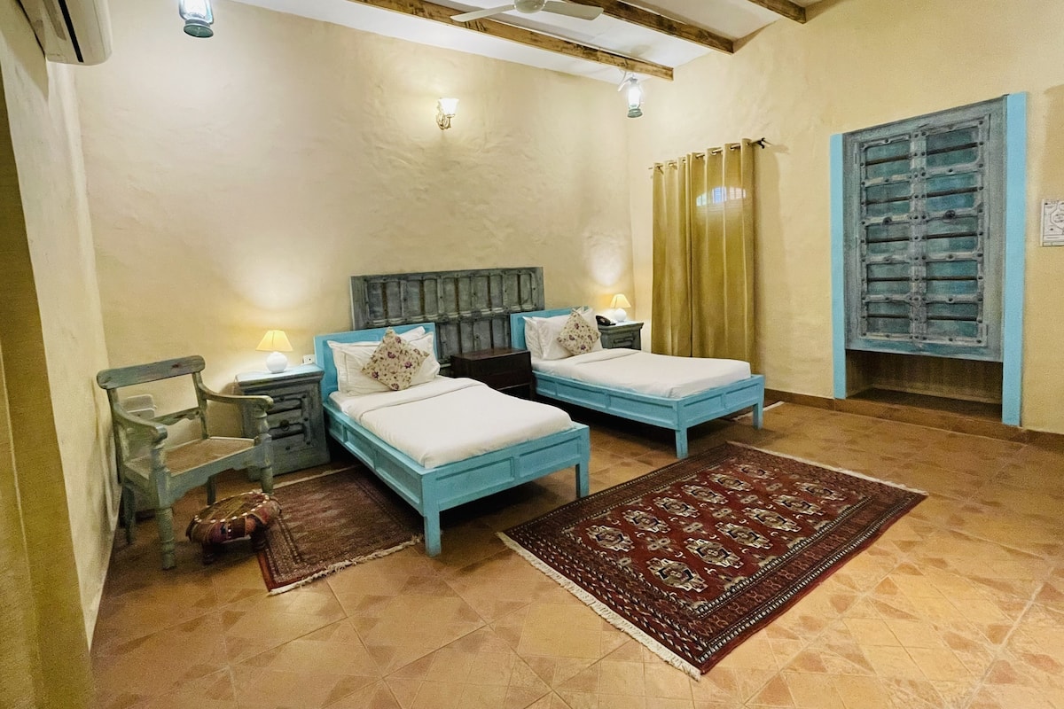A bedroom with two beds, a wooden headboard, a nightstand, a lamp, a window with curtains, and a patterned rug.