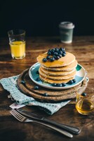 Una pila de tortitas con arándanos y almíbar sobre un plato, con un vaso de jugo y una taza al fondo.
