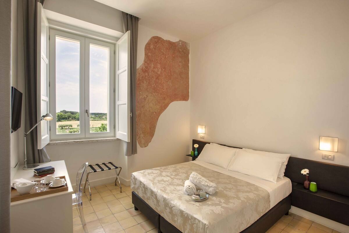 Une chambre à coucher comprenant un lit, des tables de chevet, un bureau, une chaise, une fenêtre et une vue sur la verdure à l’extérieur.
