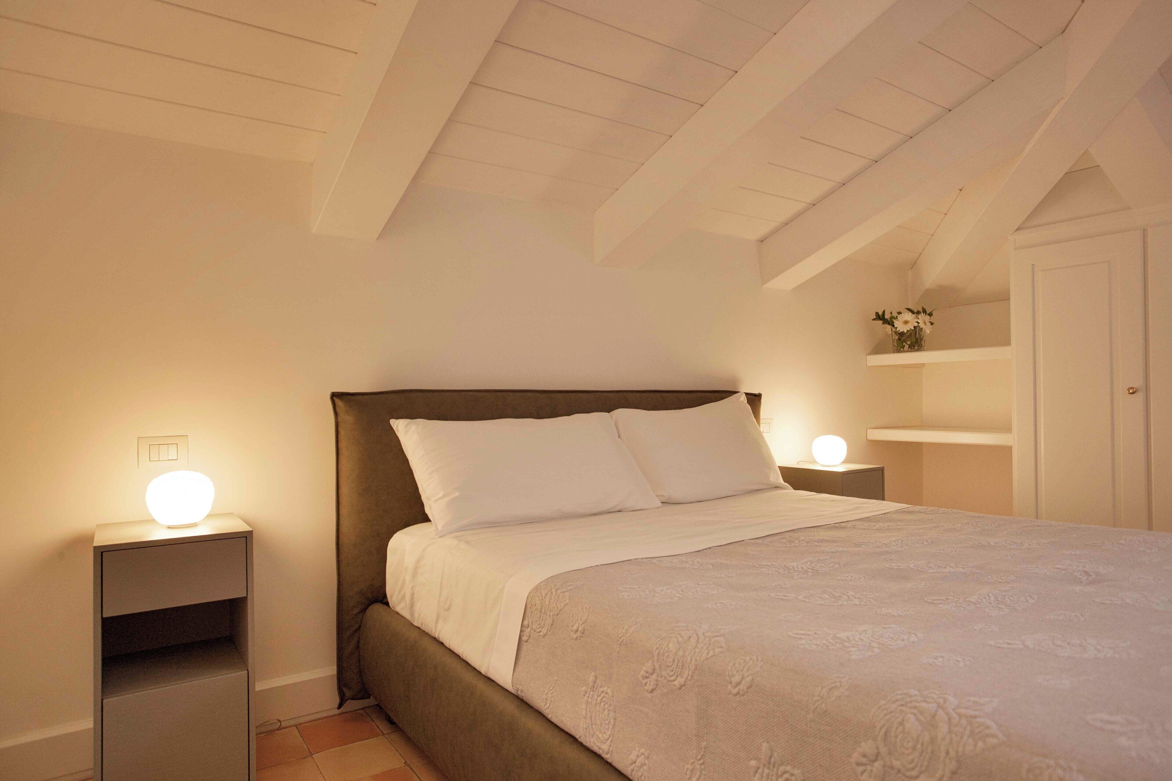 A bedroom with a bed, bedside tables, a nightstand, a shelf with flowers, and a door.