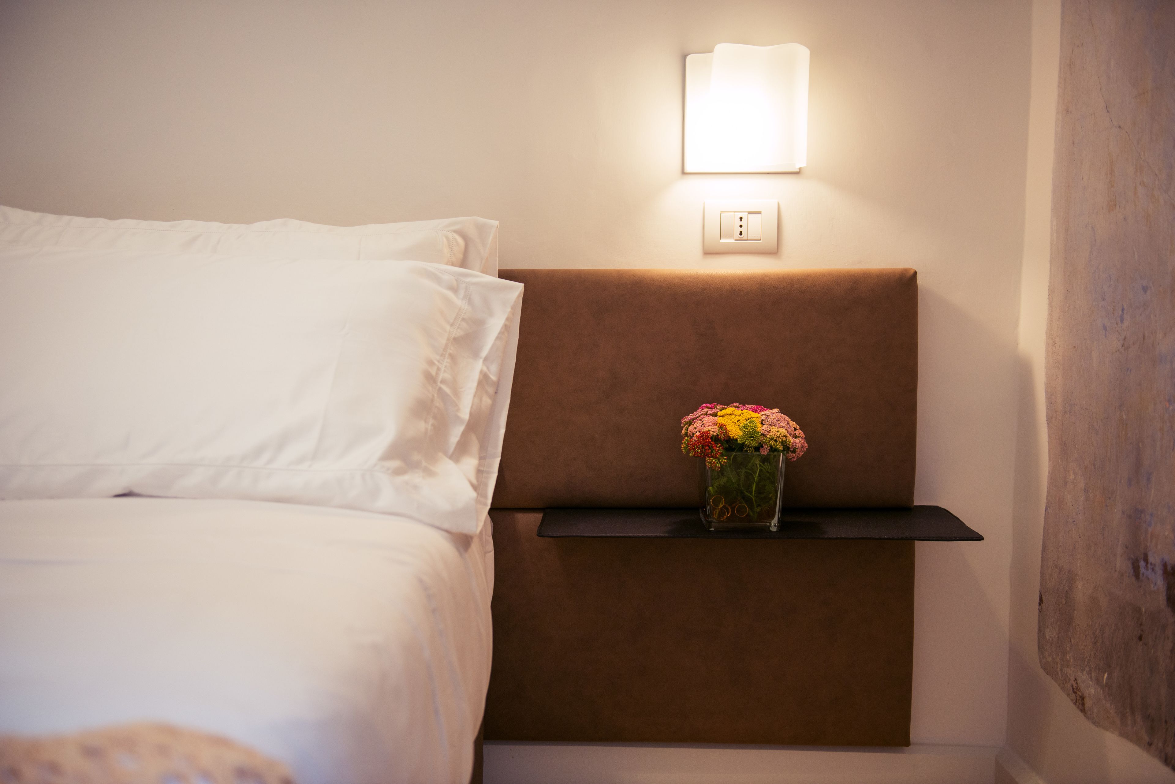 Un lit bien fait, avec du linge de lit blanc, une table de chevet ornée d’un vase de fleurs et un luminaire fixé au mur.