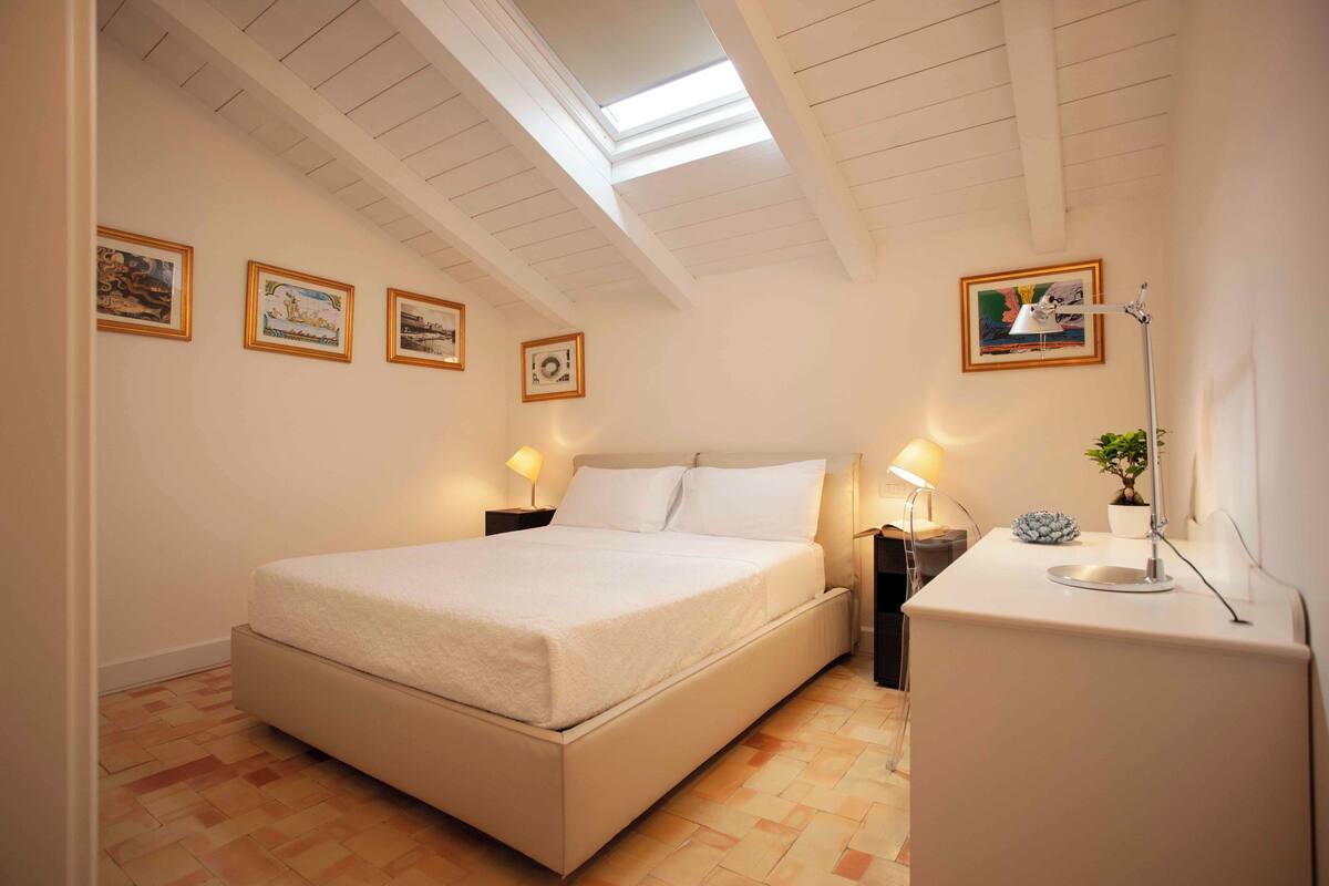 Une chambre à coucher avec un lit, des tables de chevet, un bureau, une lampe et des tableaux encadrés au mur.