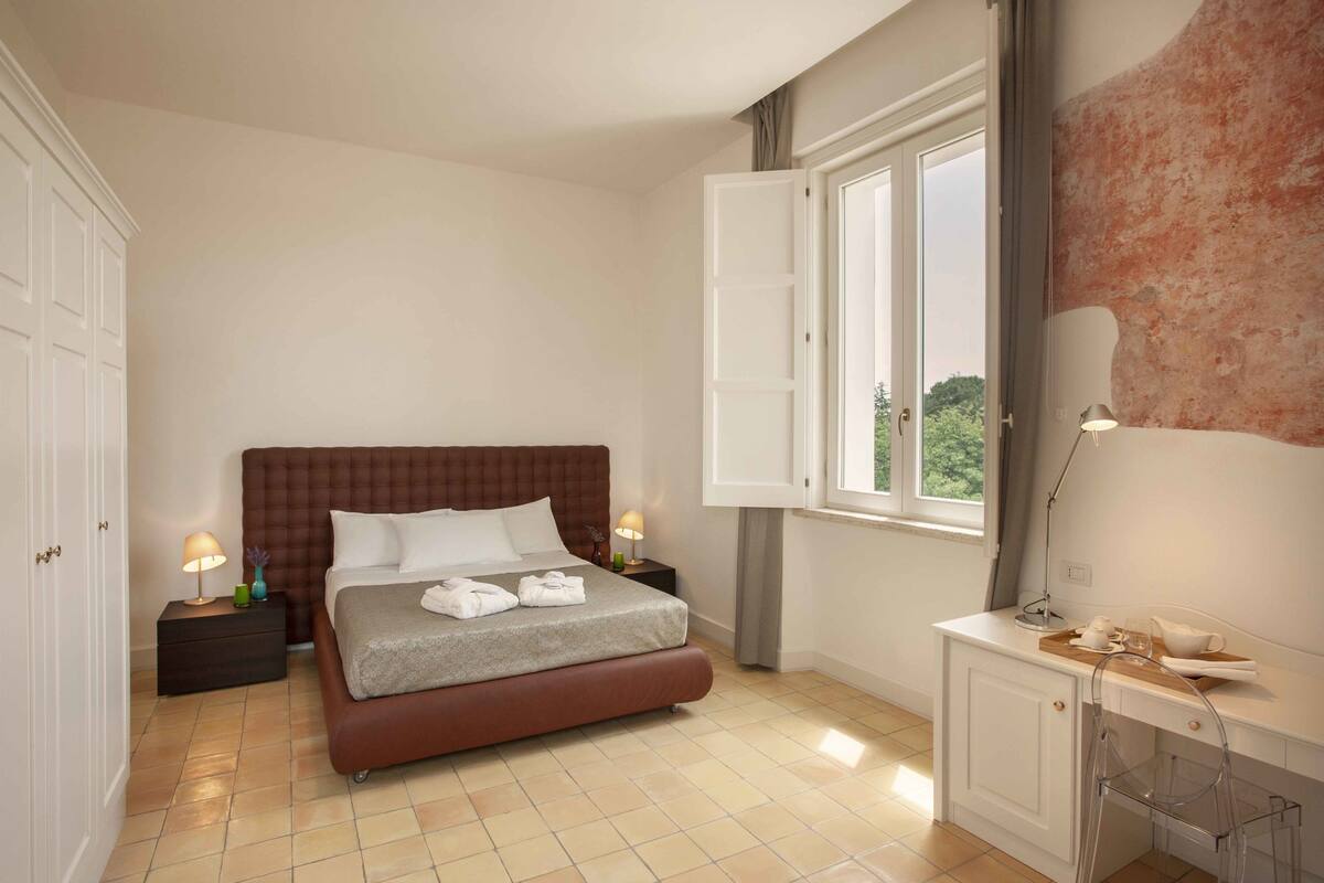 Une chambre à coucher avec un grand lit, une table de chevet avec une lampe, une fenêtre avec des rideaux et un bureau avec une chaise.