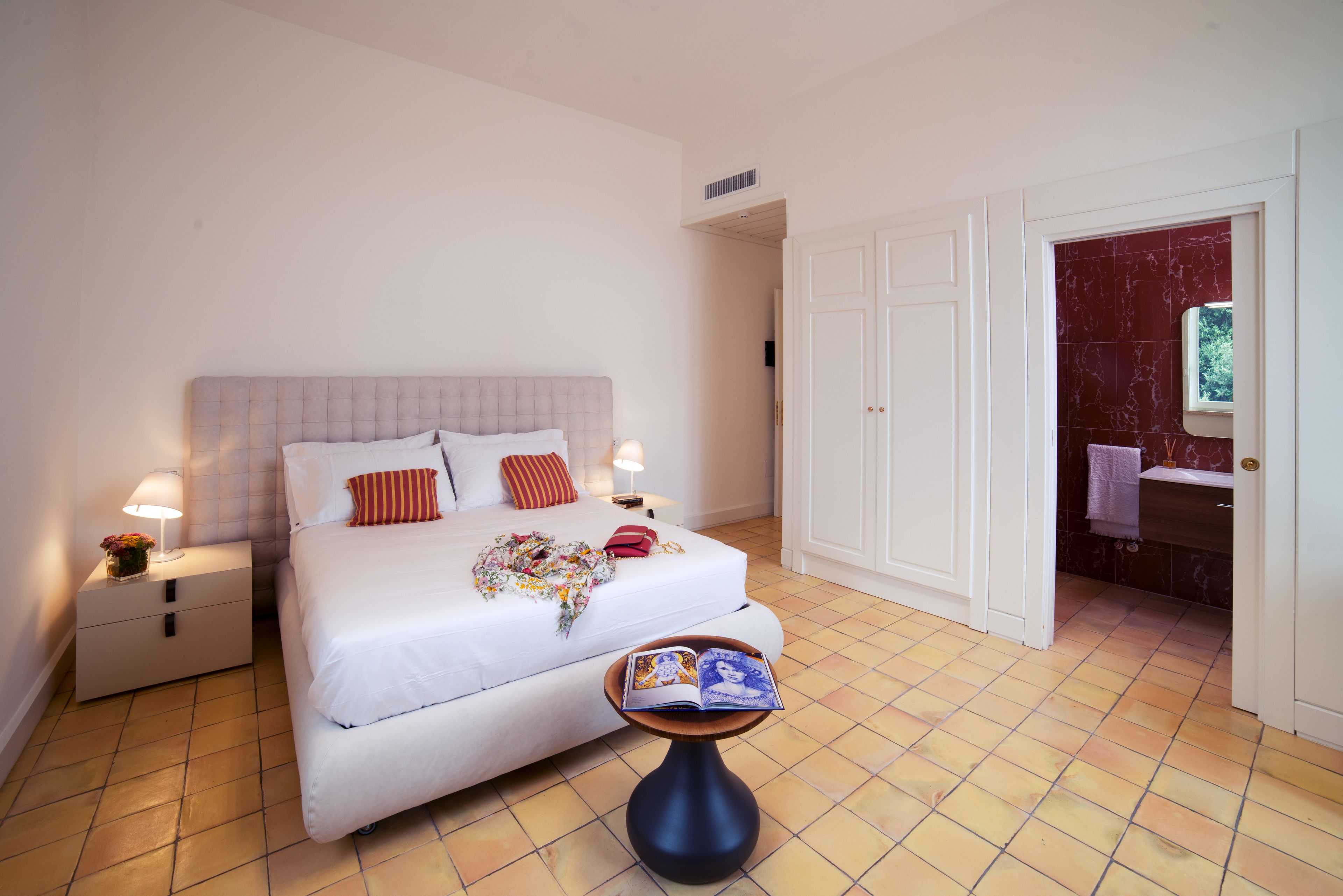 Une chambre à coucher bien rangée, avec un lit, des lampes de chevet, une petite table sur laquelle se trouve un livre, et une salle de bain visible à travers une porte ouverte.