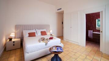 Une chambre à coucher bien rangée, avec un lit, des lampes de chevet, une petite table sur laquelle se trouve un livre, et une salle de bain visible à travers une porte ouverte.