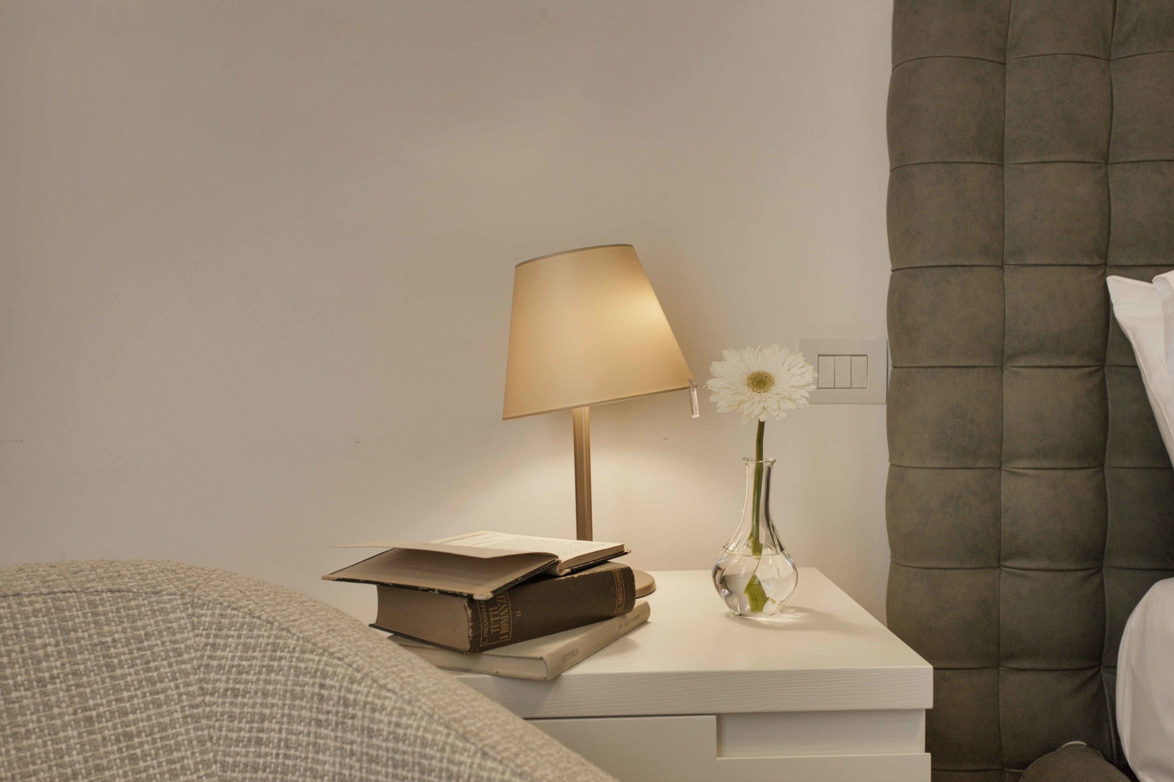 A neatly arranged bedroom with a bedside table featuring a lamp, a vase with a flower, and a stack of books.