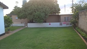 A residential backyard with a well-maintained lawn, a large tree, and a white wall with a window. - Mambilima Furnished Guest Wing (Lusaka)