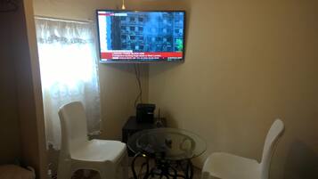 Una sala de estar con un televisor montado en la pared mostrando noticias, dos sillas blancas, una mesa de vidrio y un pequeño modelo de la Torre Eiffel sobre la mesa.
