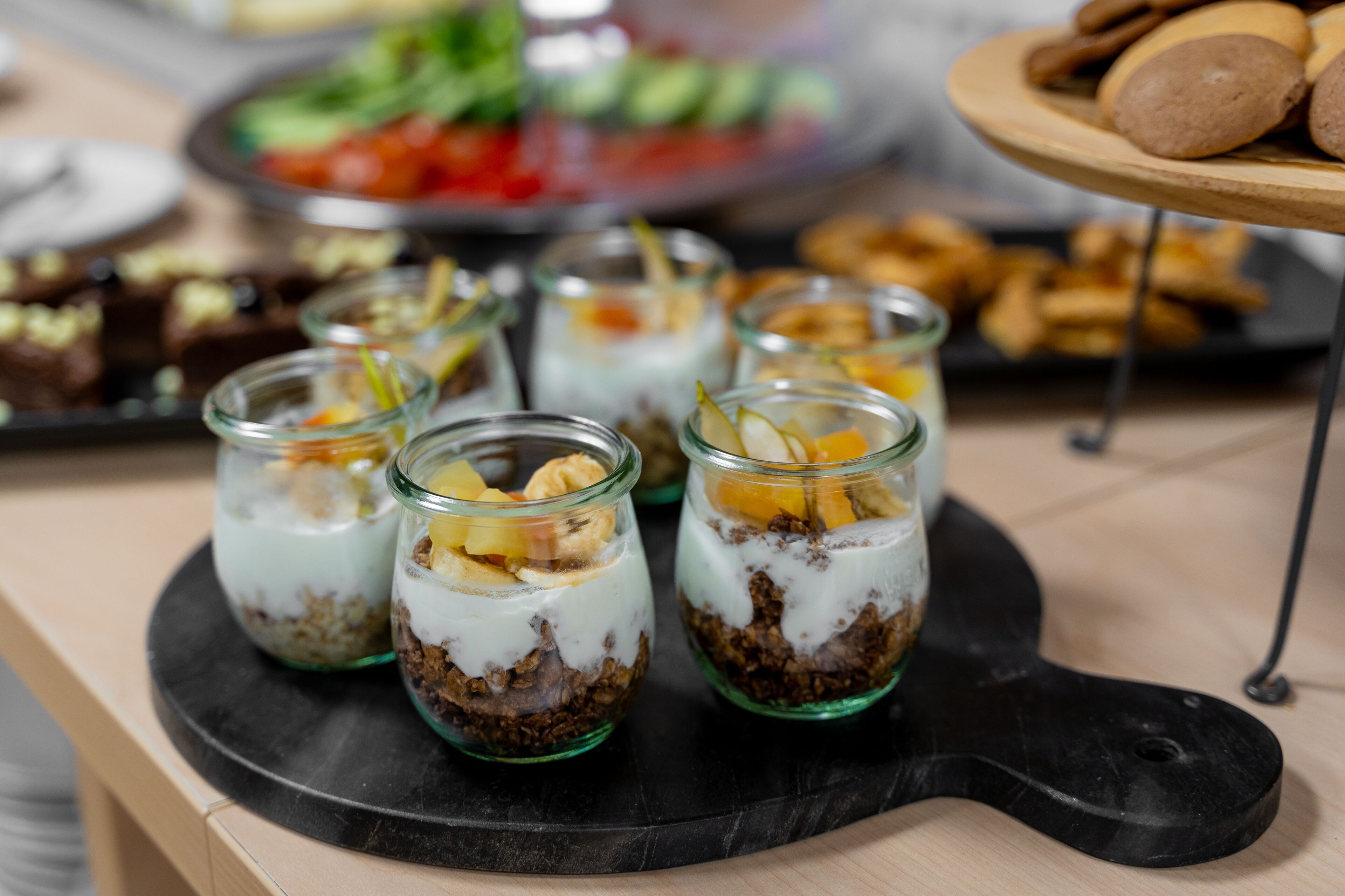 Un plateau avec six bocaux en verre contenant des aliments disposés en couches, une assiette de biscuits et une salade en arrière-plan.