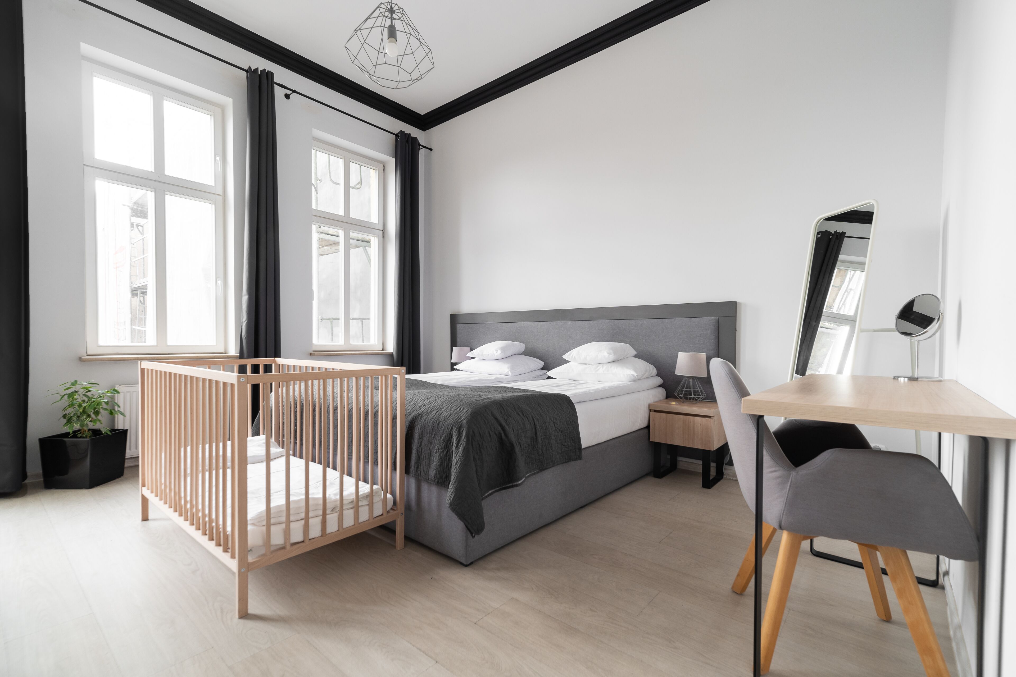 Una camera da letto moderna con una culla, un letto con testiera grigia, una scrivania con una sedia e un'ampia finestra con tende.