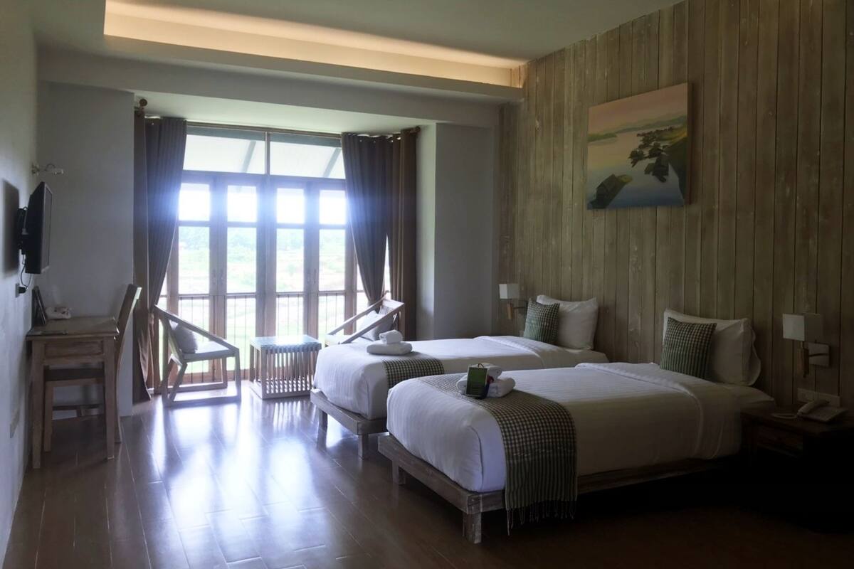 A hotel room with two beds, a wooden wall, a painting, and a view through large windows.