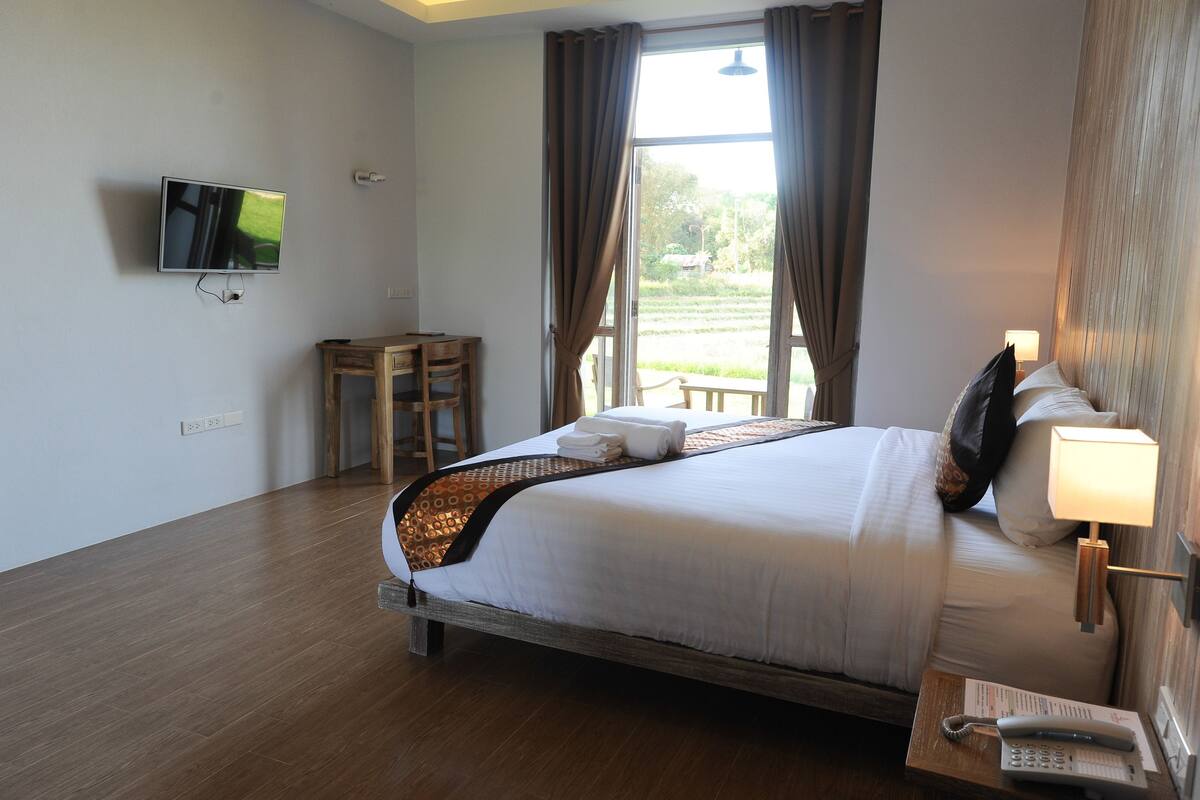 A hotel room with a bed, a TV mounted on the wall, a bedside table with a lamp, and a window with curtains.