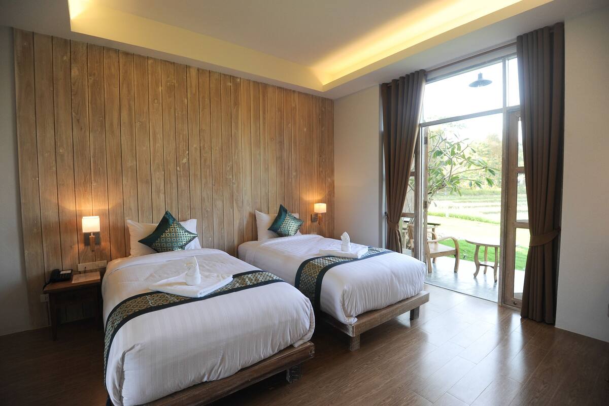 A hotel room with two beds, wooden panel walls, and a view of a garden through a glass door.