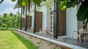 A modern outdoor seating area with wooden chairs and tables, a concrete floor, and a view of a lush green garden. - Nansripanna Resort (Nan)