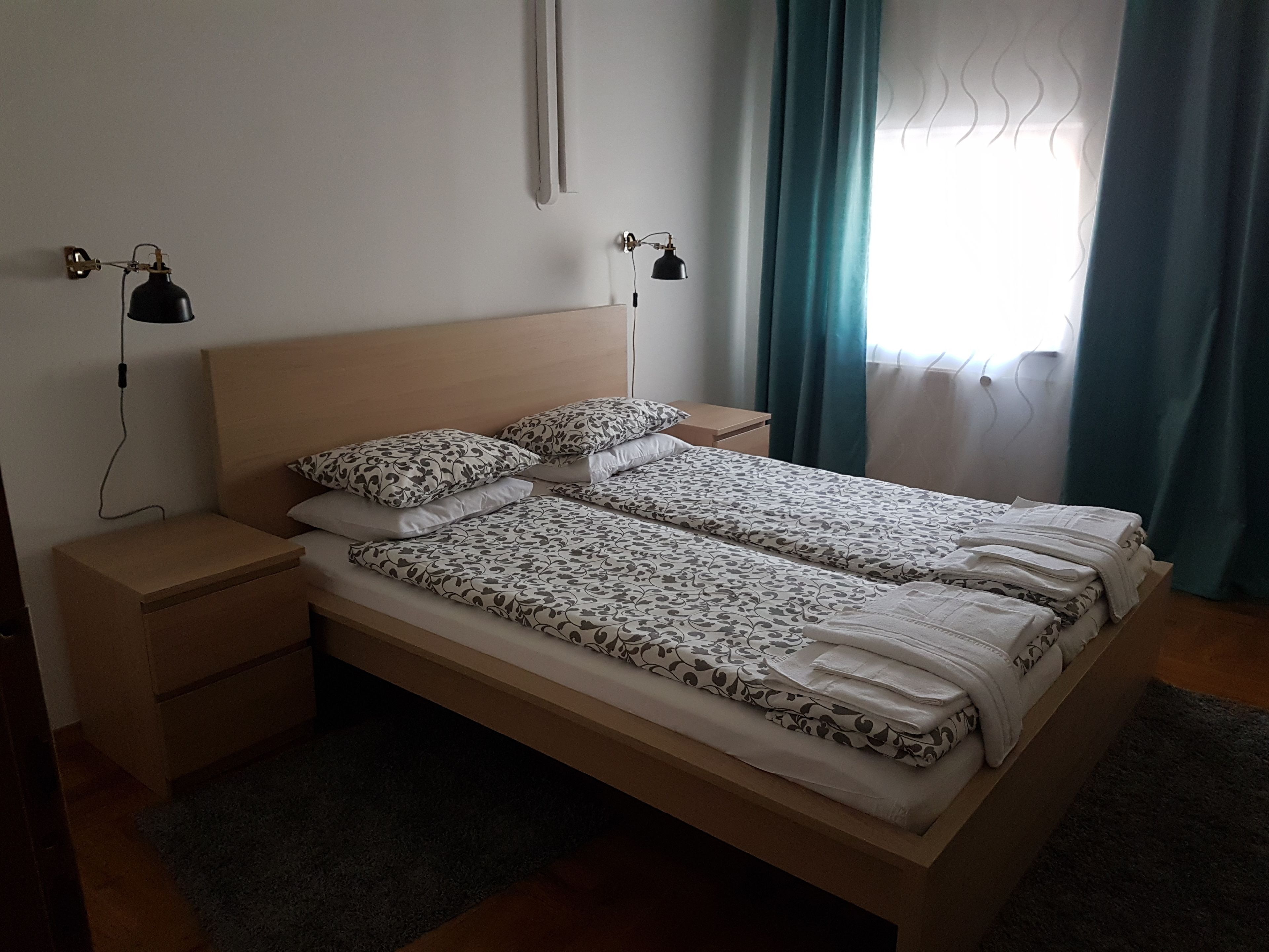 A double bed with patterned bedding, two bedside tables, a nightstand, a wall-mounted lamp, and a window with curtains.