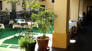 Un espace extérieur aménagé, abrité et doté de tables et de chaises, de plantes en pot, et un bâtiment avec un toit en tuiles en arrière-plan.