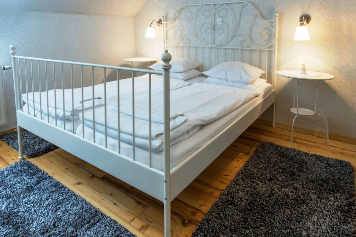 Una cama de metal blanco en una habitación con suelo de madera y dos mesitas de noche con lámparas.