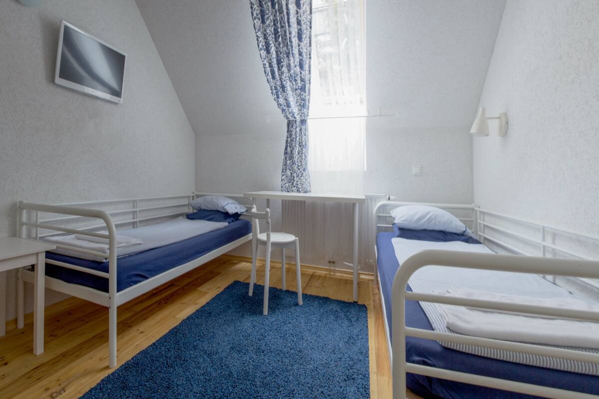 Habitación tipo dormitorio con dos camas individuales, una mesita pequeña, una silla y un televisor montado en la pared.