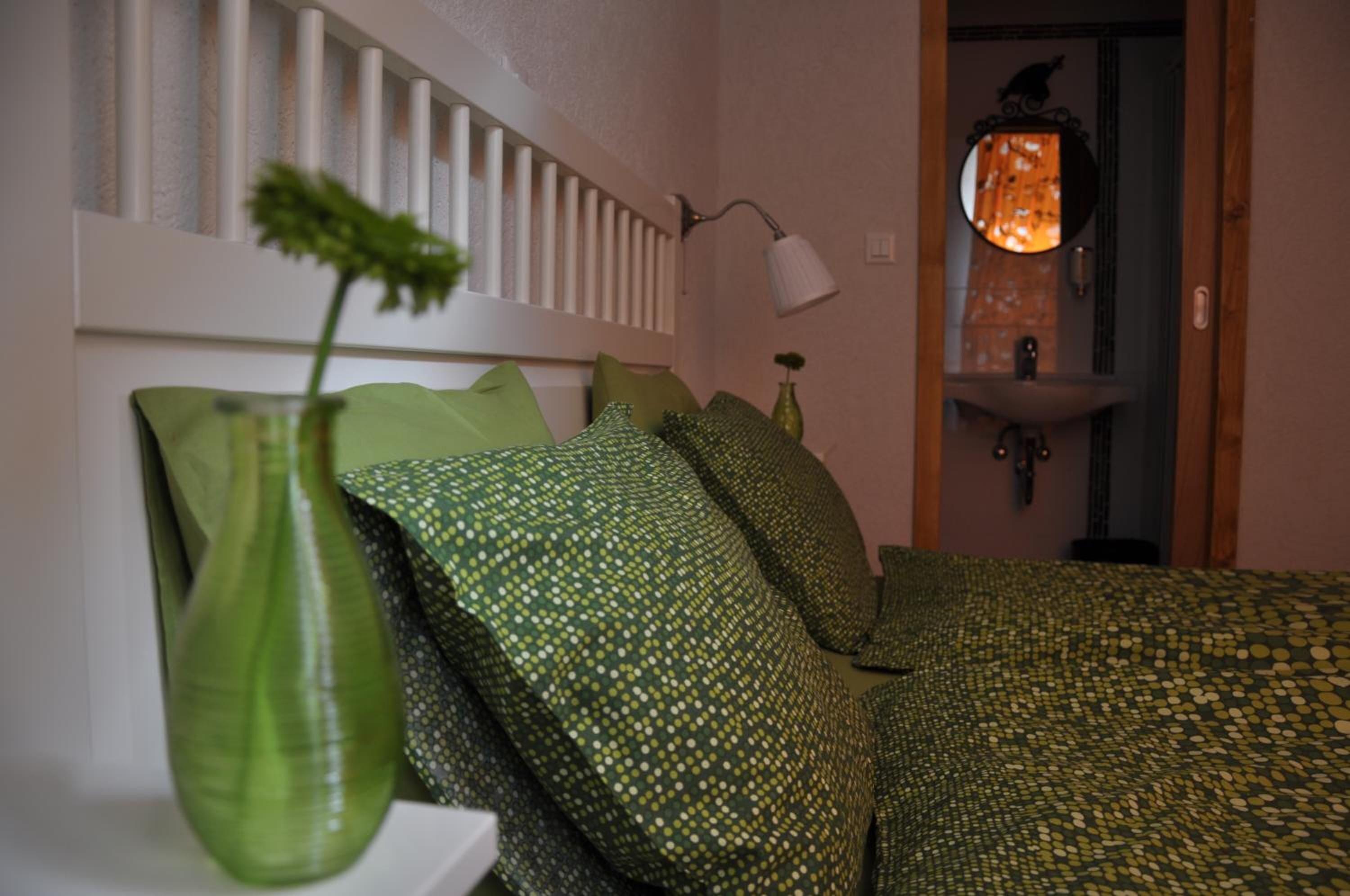 A bedroom with a bed, pillows, a bedside table with a vase, a lamp, and a view of a bathroom with a sink and mirror.