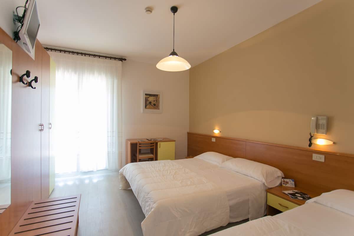 A hotel room with two beds, a desk, a chair, a small table, a lamp, a telephone, and a window with curtains.
