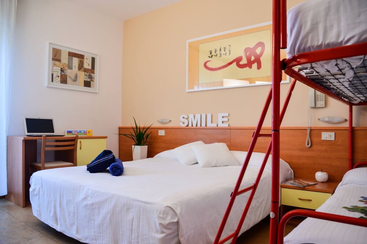 A hotel room with a bed, a desk with a laptop, a plant, and a wall with a 'SMILE' sign.
