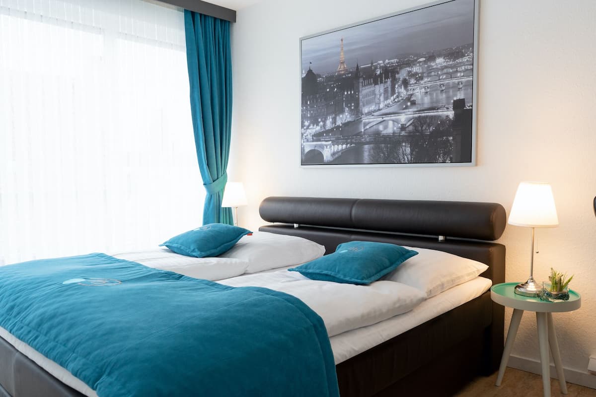 Ein Hotelzimmer mit einem Bett, zwei Kissen, einem Nachttisch mit Lampe und einem gerahmten Stadtbild an der Wand.