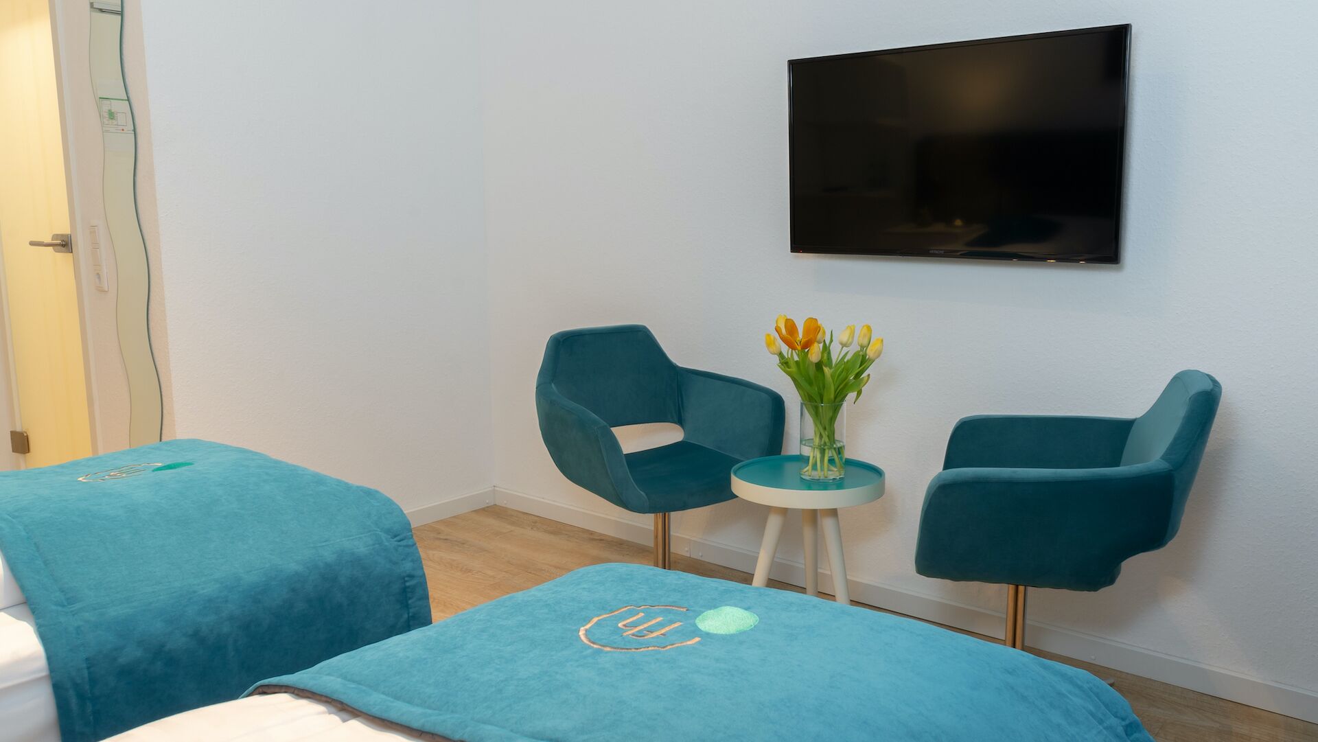 A hotel room with two teal upholstered chairs, a small round table with a vase of flowers, and a flat-screen TV mounted on the wall.
