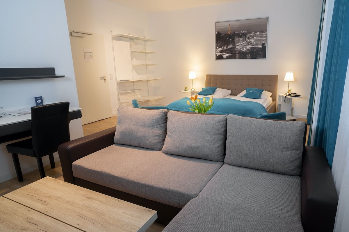 Ein modernes Hotelzimmer mit einer Couch, einem kleinen Schreibtisch, einem Bett mit blauer Bettwäsche und Blick auf die Stadt.