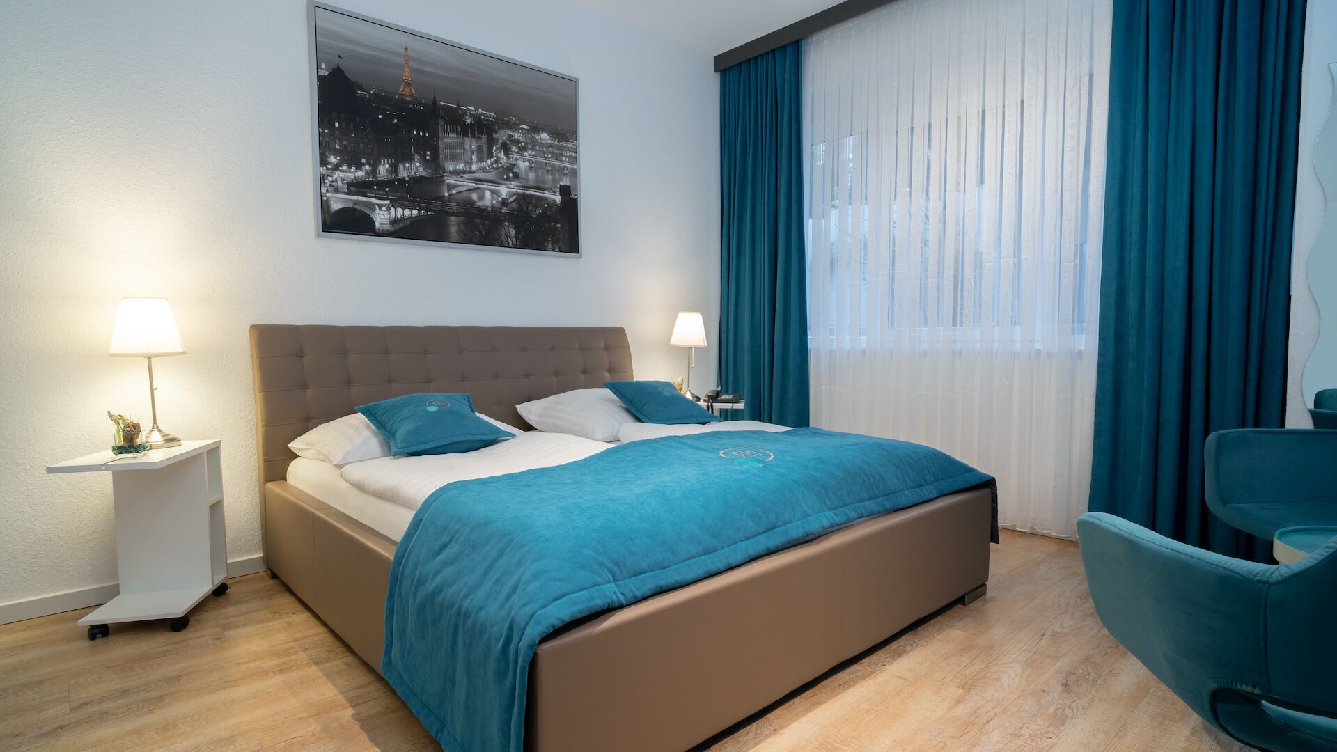 Una habitación de hotel con una cama, dos mesitas de noche, una silla, un ventanal con cortinas y vistas a la ciudad.