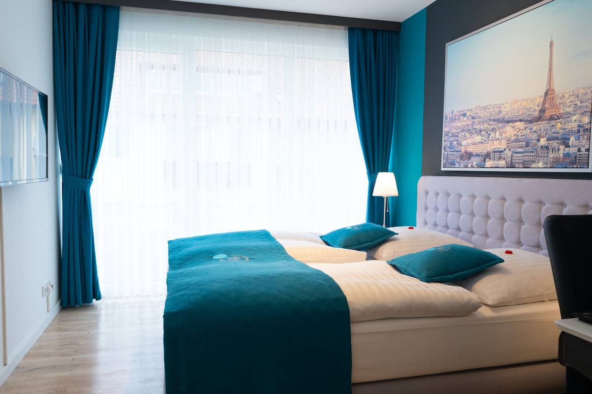 Ein Hotelzimmer mit Bett, Nachttisch, Lampe und einem großen Fenster mit Vorhängen.