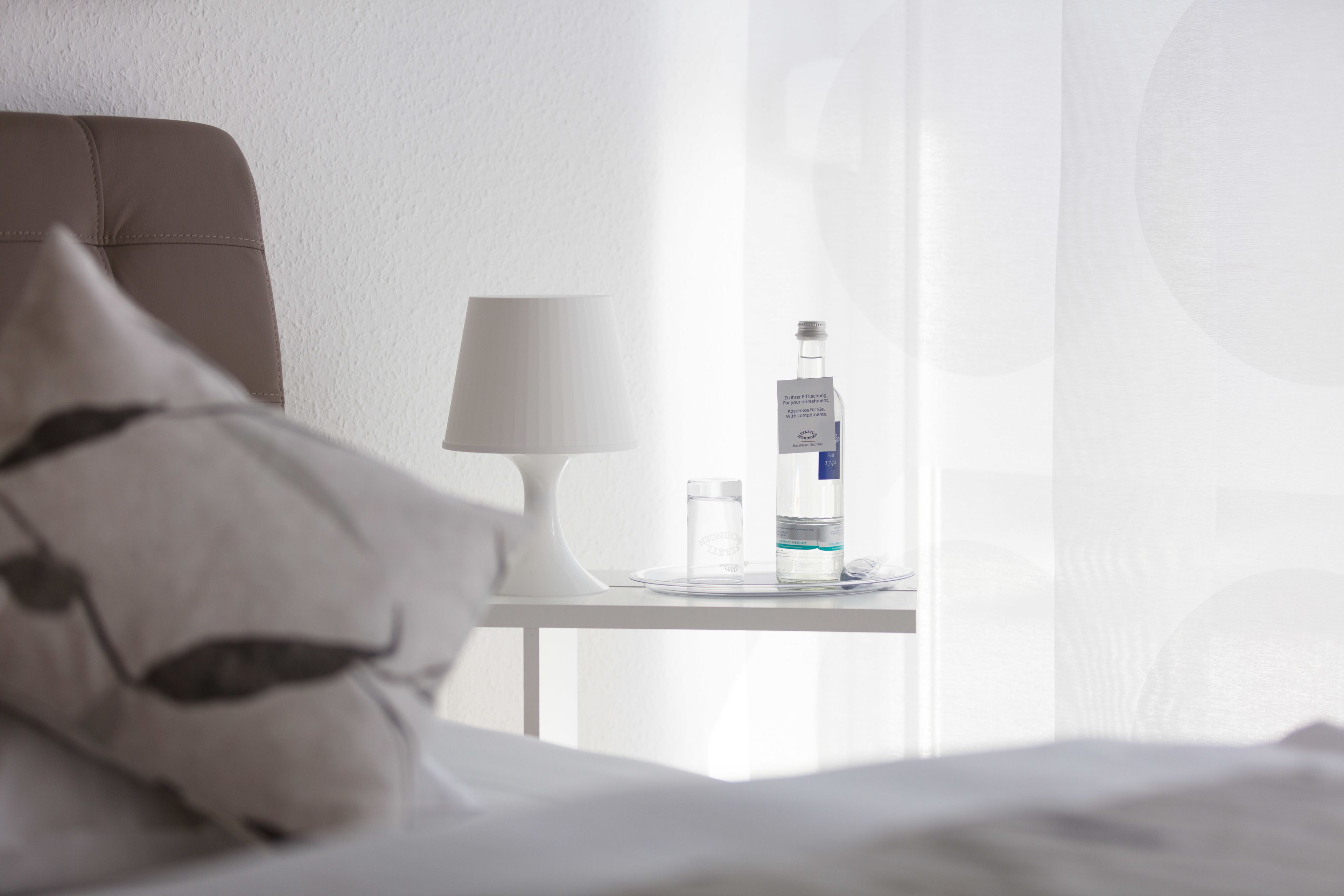 Ett hotellrum med en säng, ett nattduksbord, en lampa och en flaska vatten.
