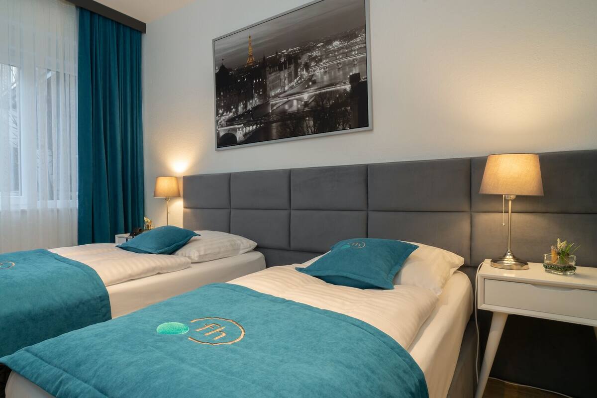 Ein Hotelzimmer mit zwei Betten, einem großen Fenster mit durchsichtigen Vorhängen, einem Stadtbild an der Wand und einem Nachttisch mit einer Lampe.