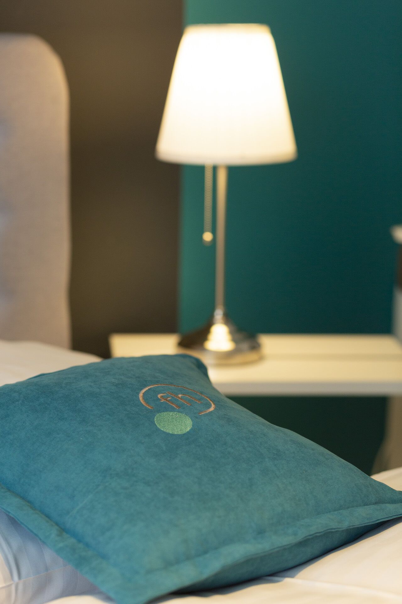 Una cama bien hecha con un almohán color turquesa y uno blanco. En el fondo, se ve una mesita con una lámpara.