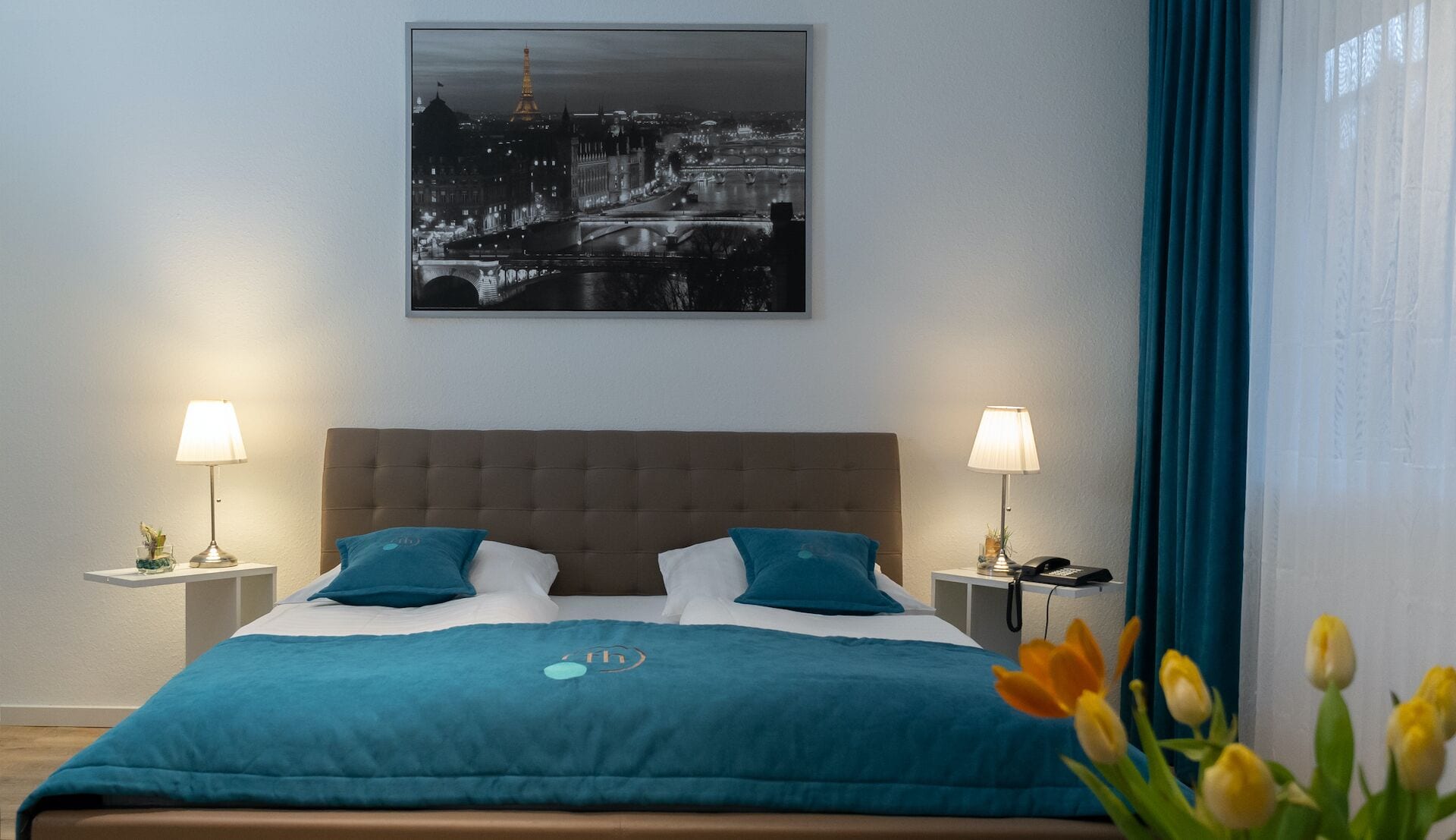 A hotel room with a large bed, two bedside lamps, a nightstand with a phone, and a framed cityscape picture on the wall.
