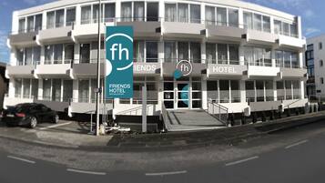 Ein modernes Gebäude mit einem Schild des Friends Hotel, das einen zentralen Eingang mit einer Rampe, mehrere Balkone und große Fenster aufweist.