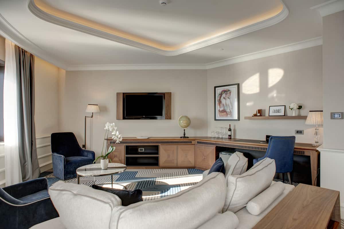 A modern hotel room with a sofa, armchairs, a coffee table, a desk, and a flat-screen TV.