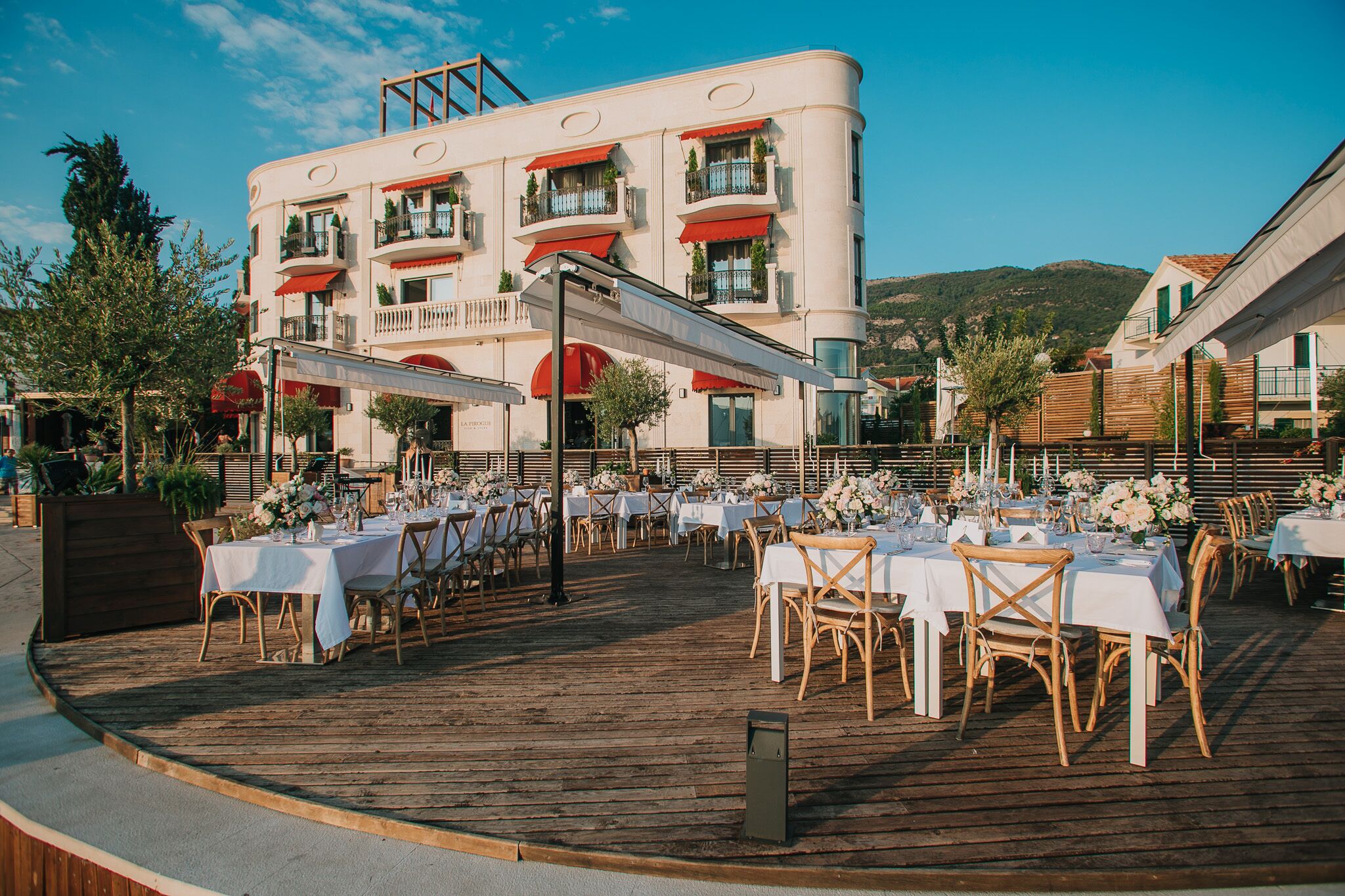 Una terraza con varias mesas y sillas, un edificio con balcones y toldos rojos, y montañas al fondo.