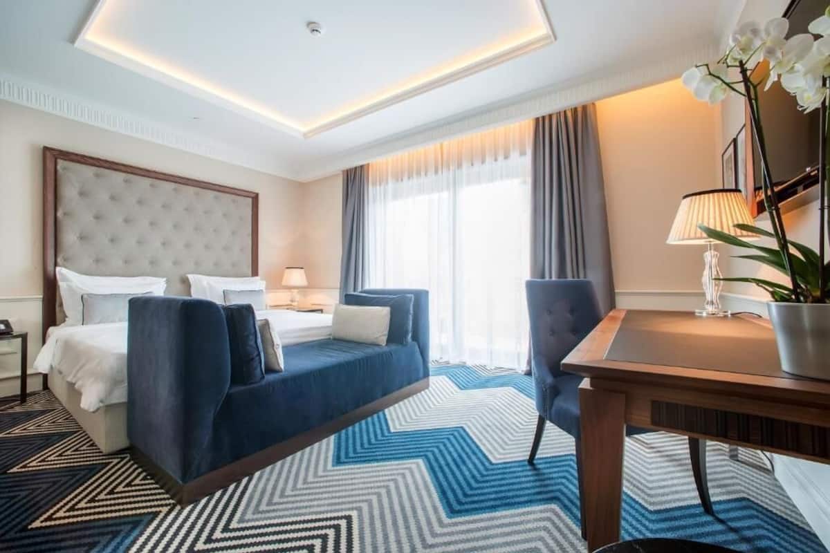 A hotel room with a bed, a desk, a chair, a lamp, and a potted plant.