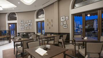 A hotel dining area with multiple tables and chairs, framed pictures on the walls, and a view through the windows.