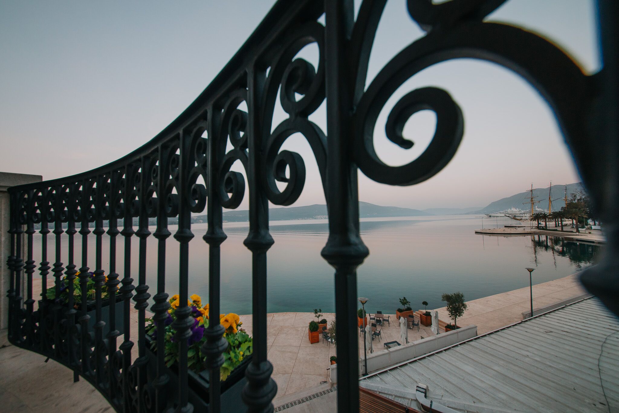 A balcony with wrought iron railings overlooking a calm body of water and distant mountains.