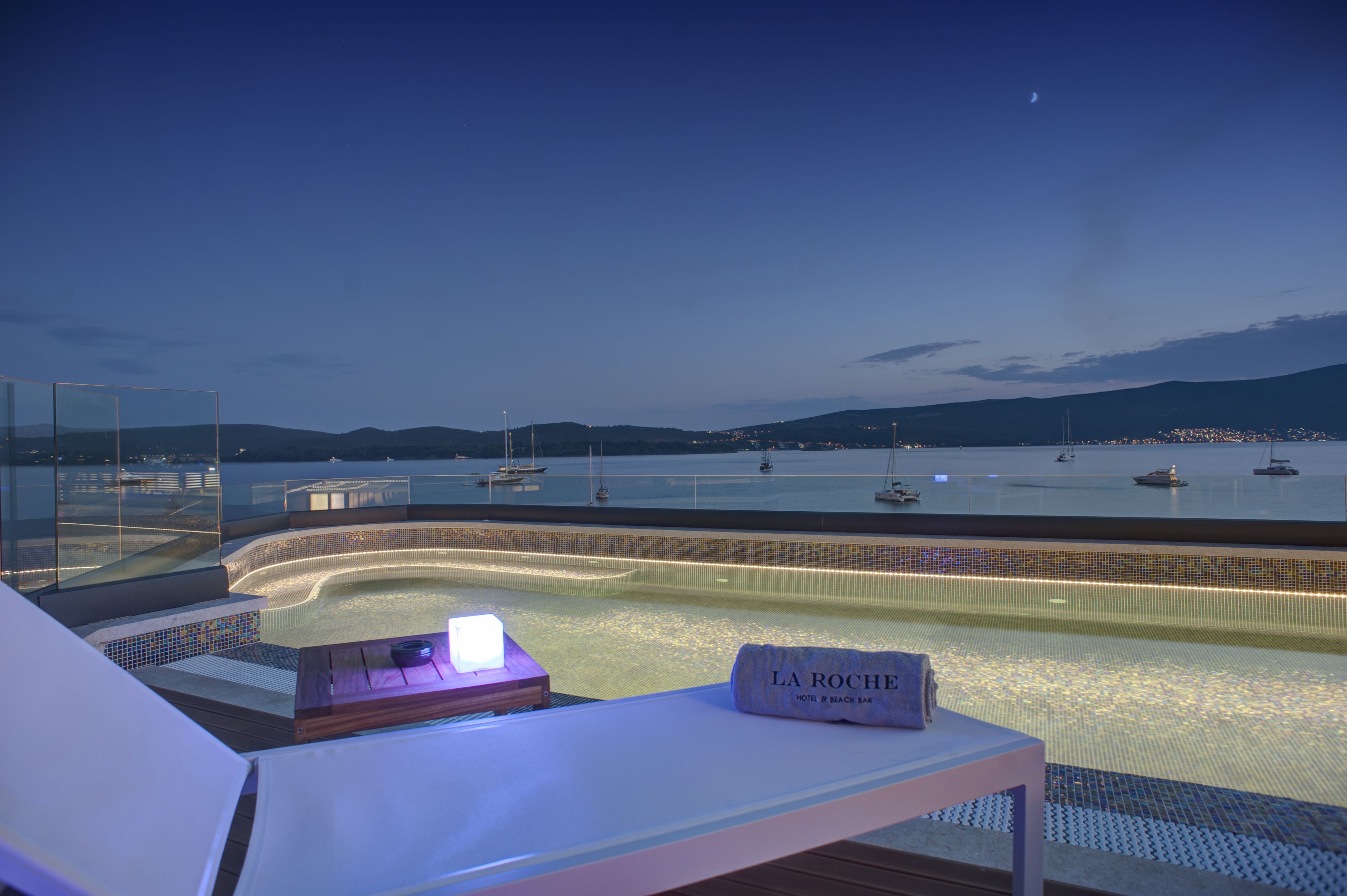 Uma área de descanso à beira da piscina com vista para o mar e barcos, uma espreguiçadeira e uma placa com a inscrição "LA ROCHE".