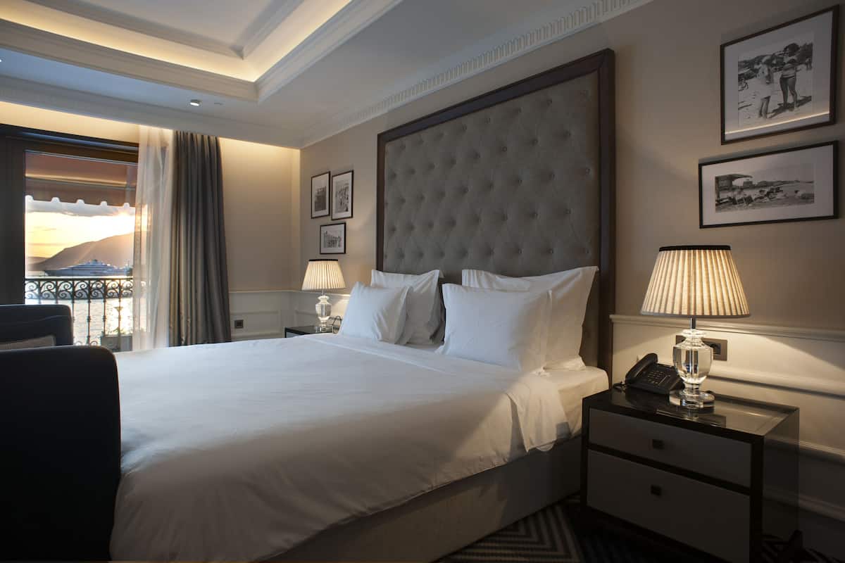 A hotel room with a large bed, a nightstand with a lamp, a chair, and a view of the sunset through the window.